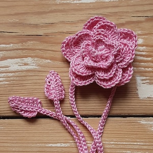 May include: A pink crocheted flower with three leaves attached to the stem. The flower is made of multiple layers of crocheted petals.