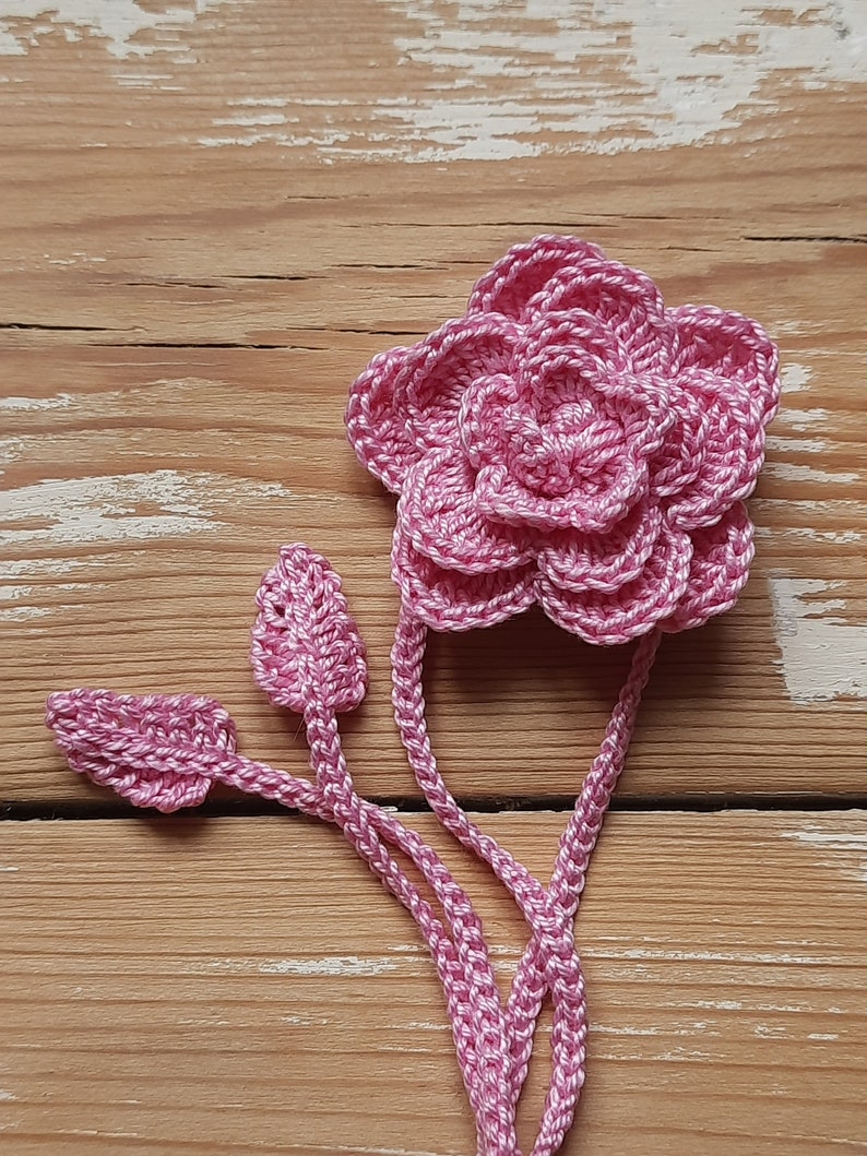 May include: A pink crocheted flower with three leaves attached to the stem. The flower is made of multiple layers of crocheted petals.