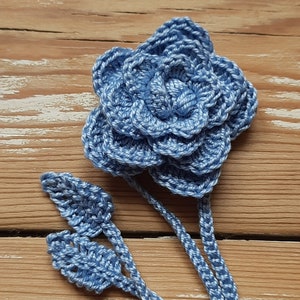 May include: A crocheted blue flower with a stem and leaves. The flower is made of multiple layers of crocheted petals.
