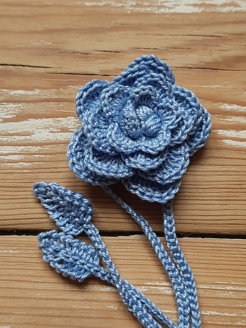May include: A crocheted blue flower with a stem and leaves. The flower is made of multiple layers of crocheted petals.