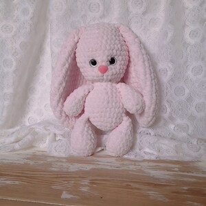 K&ouml;nnte beinhalten: Ein rosa geh&auml;keltes Hasenspielzeug mit schwarzen Augen und einer rosa Nase. Der Hase sitzt auf einer Holzoberfl&auml;che.