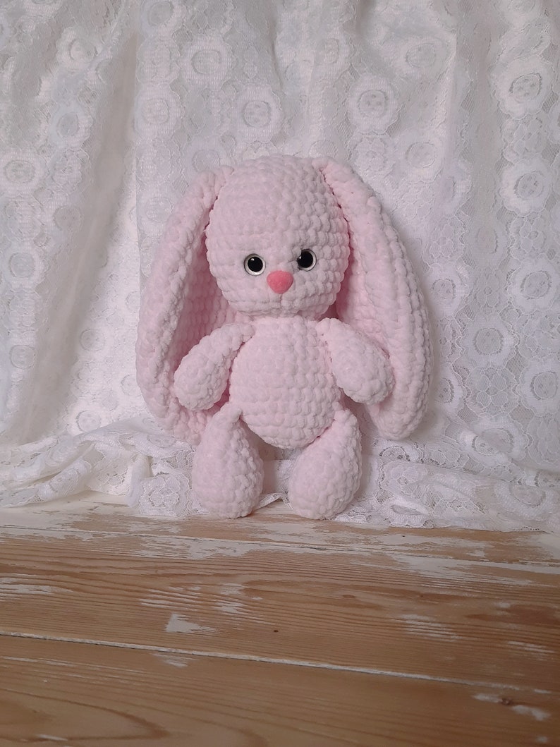K&ouml;nnte beinhalten: Ein rosa geh&auml;keltes Hasenspielzeug mit schwarzen Augen und einer rosa Nase. Der Hase sitzt auf einer Holzoberfl&auml;che.
