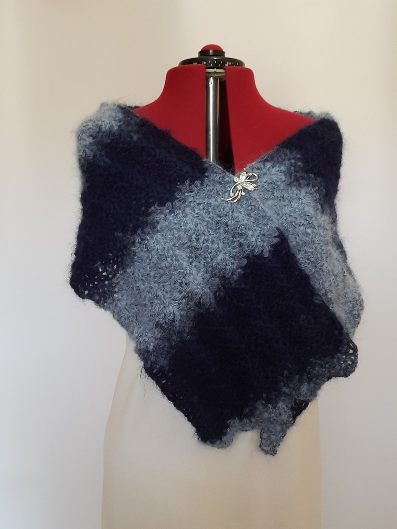 May include: A blue and gray knitted shawl with a decorative pin on a white dress form.
