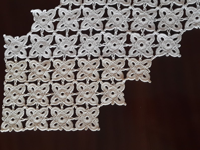 May include: White crocheted table runner with a geometric pattern of flowers. The runner has a scalloped edge and is made of delicate, intricate stitches.