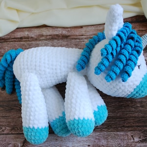 May include: A white crocheted unicorn with blue yarn for the mane, tail, and hooves. The unicorn is lying on its side on a wooden surface.