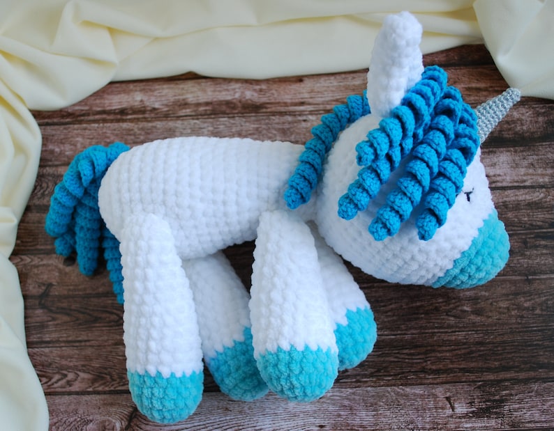 May include: A white crocheted unicorn with blue yarn for the mane, tail, and hooves. The unicorn is lying on its side on a wooden surface.