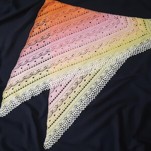 May include: A handmade, triangular shawl in a gradient of colors, transitioning from orange to pink and finally to yellow. The shawl features a delicate, openwork design with a scalloped, white lace border. The shawl is displayed on a dark background.