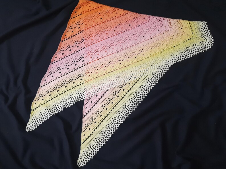 May include: A handmade, triangular shawl in a gradient of colors, transitioning from orange to pink and finally to yellow. The shawl features a delicate, openwork design with a scalloped, white lace border. The shawl is displayed on a dark background.