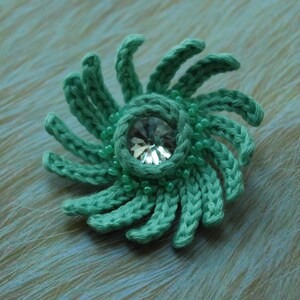 May include: A teal crocheted flower with a clear rhinestone in the centre. The flower is surrounded by small teal beads.