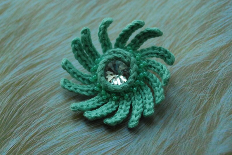 May include: A teal crocheted flower with a clear rhinestone in the centre. The flower is surrounded by small teal beads.