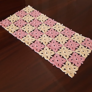 May include: A crocheted table runner featuring a pink and cream floral design. The runner is crafted from fine yarn and has a rectangular shape.