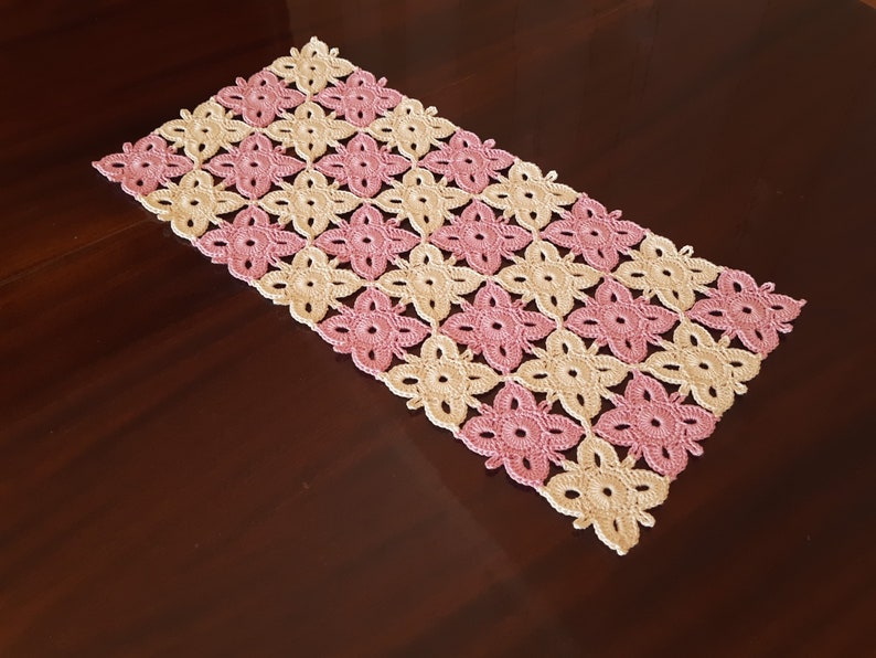 May include: A crocheted table runner featuring a pink and cream floral design. The runner is crafted from fine yarn and has a rectangular shape.