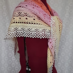 May include: A handmade crochet shawl in a gradient of colors, including peach, pink, and yellow. The shawl features an openwork design with a scalloped edge. The item is displayed on a red dress form.