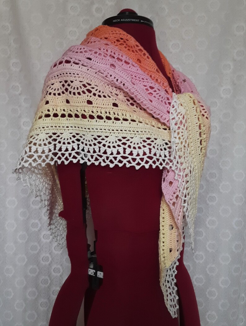 May include: A handmade crochet shawl in a gradient of colors, including peach, pink, and yellow. The shawl features an openwork design with a scalloped edge. The item is displayed on a red dress form.