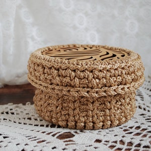Pu&ograve; includere: Una scatola di immagazzinaggio rotonda in crochet con un coperchio in legno che presenta un design decorativo tagliato al laser. La scatola &egrave; beige e il coperchio &egrave; di colore marrone chiaro.