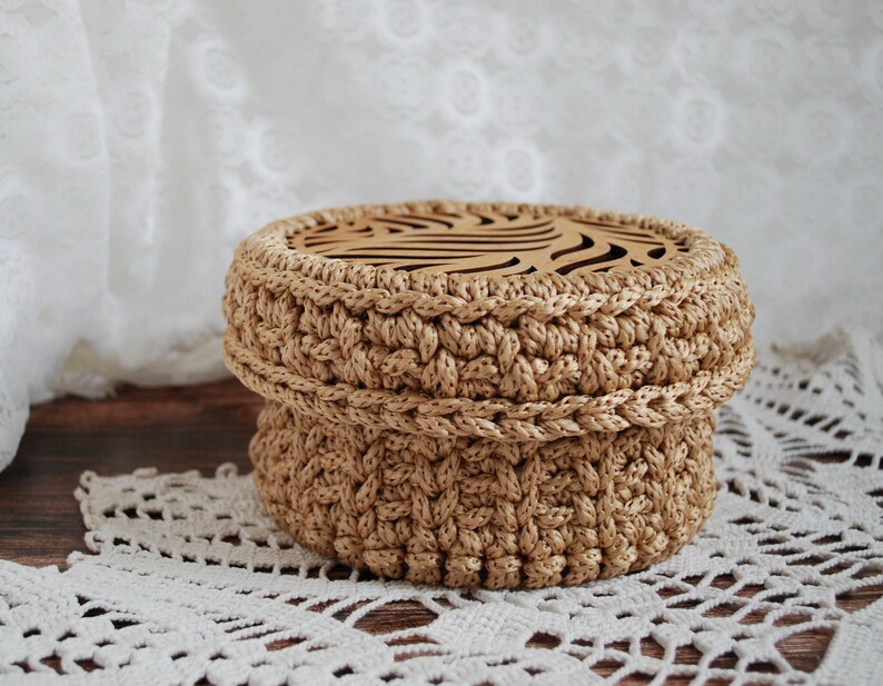 Pu&ograve; includere: Una scatola di immagazzinaggio rotonda in crochet con un coperchio in legno che presenta un design decorativo tagliato al laser. La scatola &egrave; beige e il coperchio &egrave; di colore marrone chiaro.