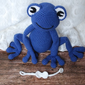 Pode incluir: Uma r&atilde; azul de croch&ecirc; com olhos brancos e pretos, sobre uma superf&iacute;cie de madeira. Uma gravata borboleta branca &eacute; colocada em frente &agrave; r&atilde;. O fundo &eacute; um tecido de renda branco.