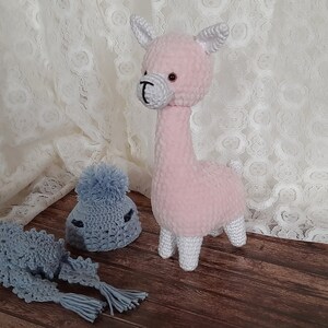 May include: A pink crocheted llama toy with white hooves and a gray crocheted hat and scarf.