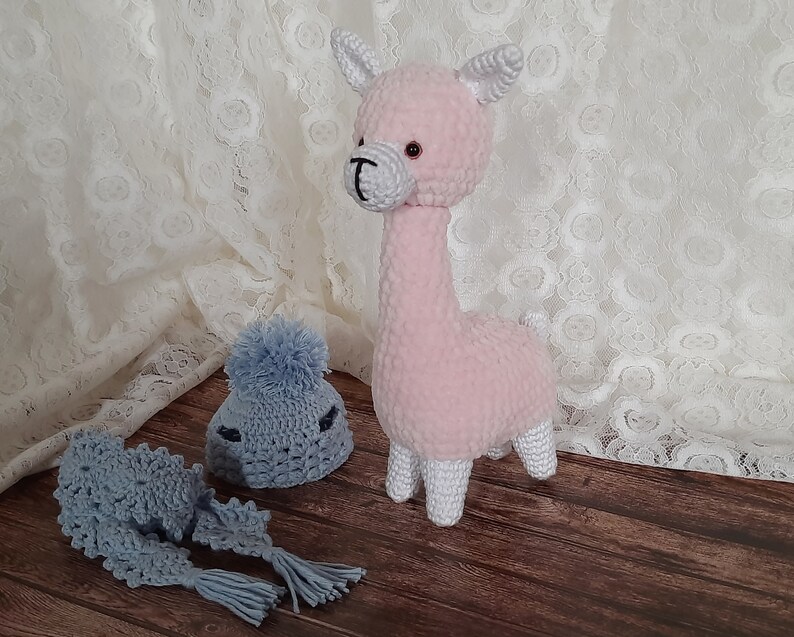 May include: A pink crocheted llama toy with white hooves and a gray crocheted hat and scarf.