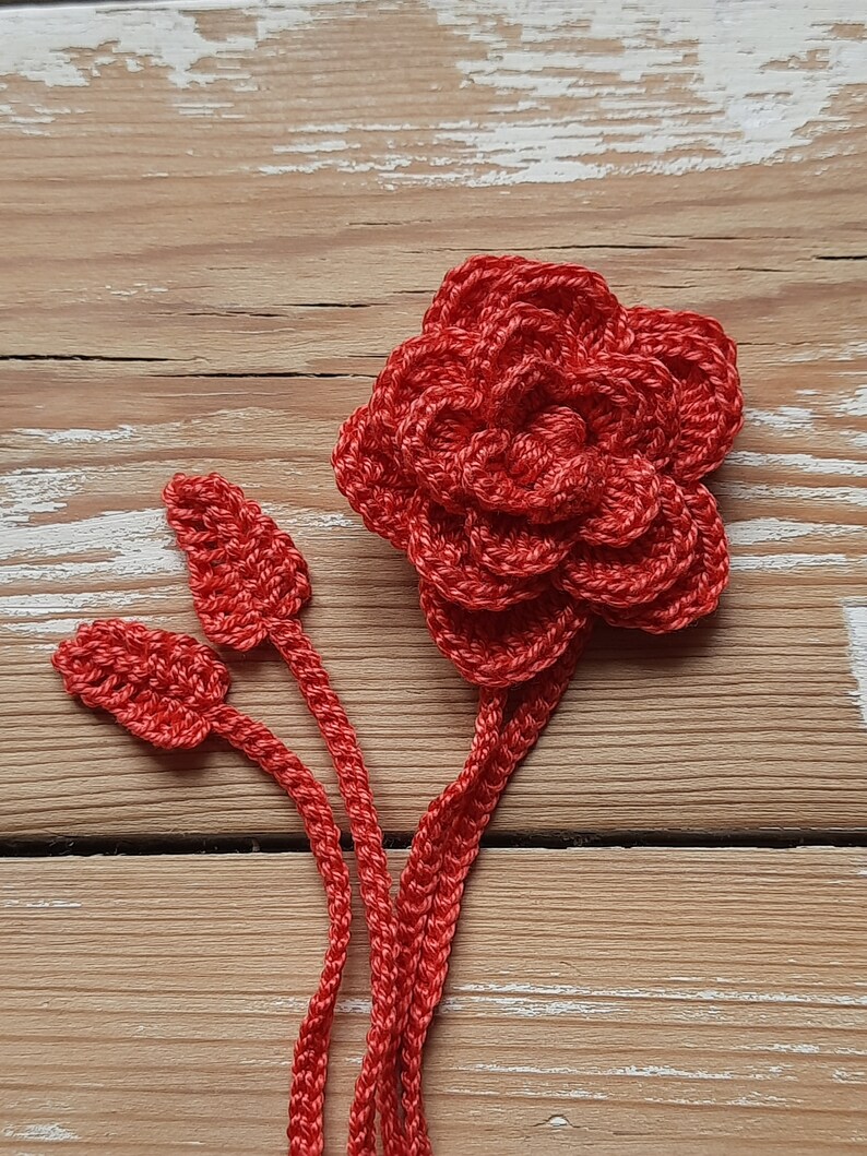 May include: A crocheted red flower with three stems and leaves. The flower is made of multiple layers of crocheted circles.