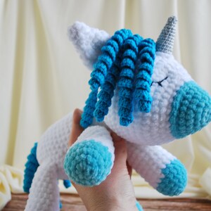 May include: A white crocheted unicorn with blue yarn for the mane and hooves. The unicorn has a silver horn and closed eyes.
