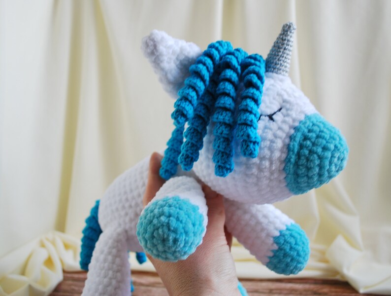 May include: A white crocheted unicorn with blue yarn for the mane and hooves. The unicorn has a silver horn and closed eyes.