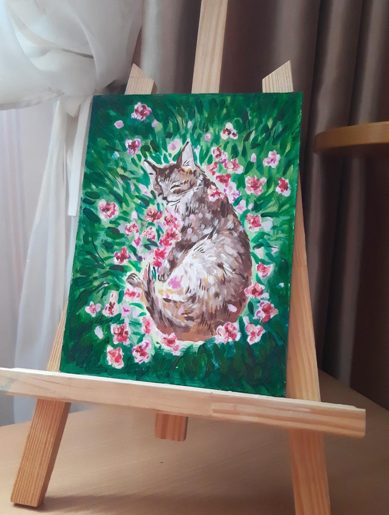 Puede incluir: Una pintura de un gato atigrado marr&oacute;n durmiendo en un campo de flores rosas. El gato est&aacute; acurrucado de lado con los ojos cerrados. Las flores est&aacute;n esparcidas alrededor del gato, y el fondo es un campo verde exuberante.