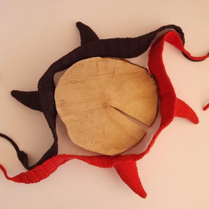 May include: Two crocheted headbands, one in solid black and one in solid red, each featuring two pointed horn-like protrusions. The headbands are arranged in a circular pattern around a central, light-coloured circular wooden slice with a visible crack.