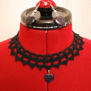 May include: A black crocheted choker necklace with a heart-shaped pendant. The pendant is made of a dark blue stone with silver glitter. The necklace is displayed on a red mannequin.