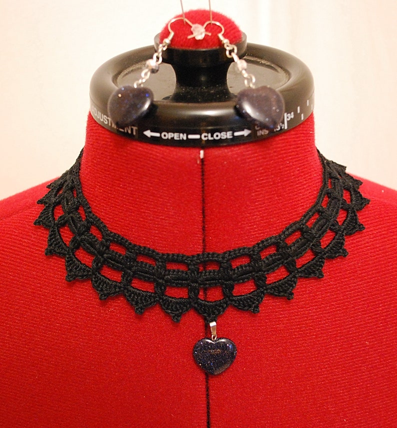 May include: A black crocheted choker necklace with a heart-shaped pendant. The pendant is made of a dark blue stone with silver glitter. The necklace is displayed on a red mannequin.