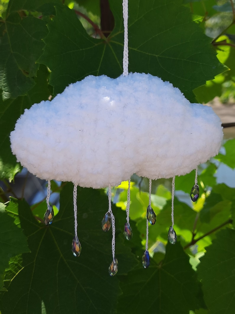Puede incluir: M&oacute;vil de nube blanca y esponjosa hecho a mano con l&aacute;grimas de cristal colgantes. La nube est&aacute; hecha de un material suave y texturizado, y los cristales iridiscentes est&aacute;n suspendidos por hilos blancos. El fondo presenta hojas verdes.