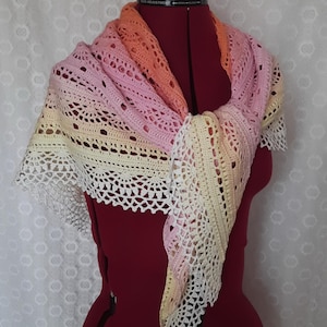 May include: A handmade crochet shawl in a gradient of colors, including pink, orange, and yellow. The shawl is draped over a red dress form, showcasing its intricate openwork design and delicate, scalloped edges. The shawl is a triangular shape.