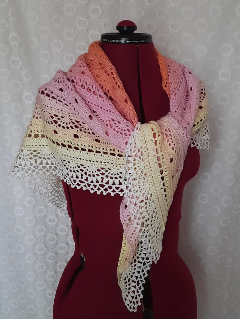 May include: A handmade crochet shawl in a gradient of colors, including pink, orange, and yellow. The shawl is draped over a red dress form, showcasing its intricate openwork design and delicate, scalloped edges. The shawl is a triangular shape.