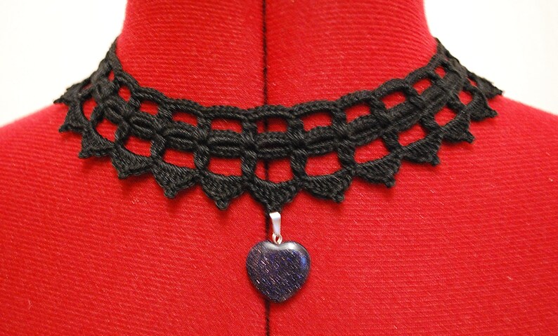 May include: A black crocheted choker necklace with a heart-shaped pendant. The pendant is made of a dark blue stone with a sparkly finish.