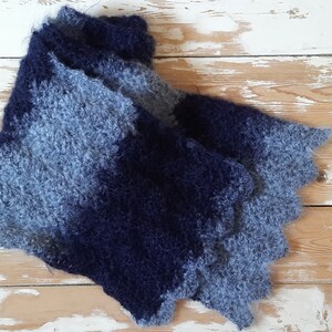May include: A blue and gray knitted scarf with a fuzzy texture. The scarf is folded in half and laid on a white wooden surface.