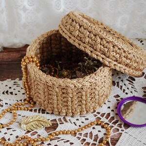 Pu&ograve; includere: Una scatola in crochet con un coperchio, realizzata in filato beige naturale. La scatola &egrave; piena di gioielli dorati e si trova su un pizzo all'uncinetto bianco. Un fermaglio a forma di piuma dorata &egrave; visibile anche sul pizzo.