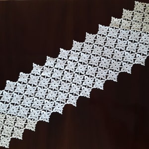 May include: White crocheted table runner with a geometric pattern of small flowers. The runner is made of delicate, lacy yarn and has a scalloped edge.