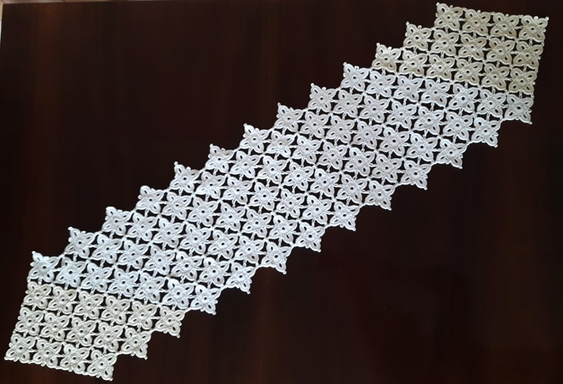 May include: White crocheted table runner with a geometric pattern of small flowers. The runner is made of delicate, lacy yarn and has a scalloped edge.