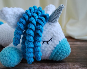 Könnte beinhalten: Ein weißes gehäkeltes Einhorn mit einer blauen Garnmähne und einem blauen Schwanz, das auf einer Holzoberfläche liegt. Das Einhorn hat ein silbernes Glitzerhorn.