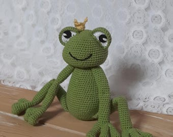 Könnte beinhalten: Ein grüner Häkel-Frosch mit einer goldenen Krone. Der Frosch hat große schwarze Augen und ein breites Lächeln. Der Frosch sitzt auf einer Holzoberfläche.