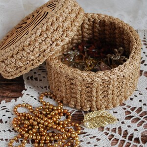 Pu&ograve; includere: Una scatola per gioielli rotonda in crochet con un coperchio. La scatola &egrave; realizzata in filato beige naturale e ha una base in legno con un motivo decorativo. La scatola &egrave; aperta e piena di gioielli, tra cui collane, orecchini e anelli.