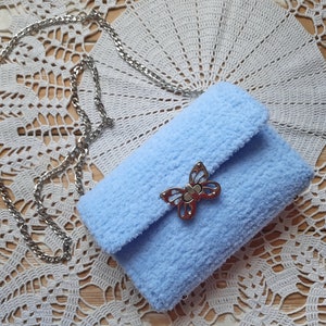 Puede incluir: Un bolso de peluche azul claro con un cierre de mariposa plateado y una correa de cadena plateada.