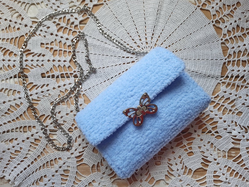 Puede incluir: Un bolso de peluche azul claro con un cierre de mariposa plateado y una correa de cadena plateada.