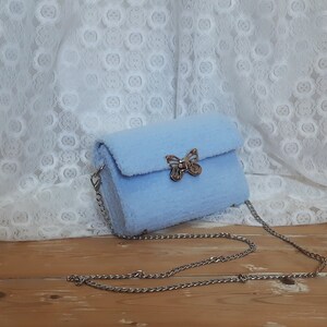 Puede incluir: Un bolso de hombro de color azul claro y peludo con un cierre de mariposa plateado y una correa de cadena plateada.