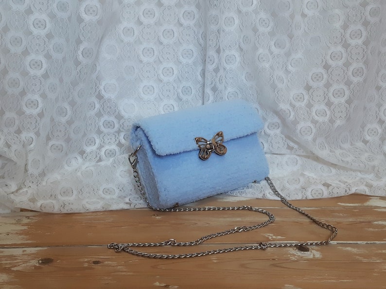 Puede incluir: Un bolso de hombro de color azul claro y peludo con un cierre de mariposa plateado y una correa de cadena plateada.