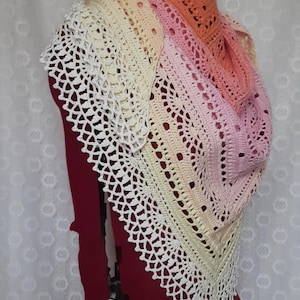 May include: A handmade crochet shawl in a gradient of colors, from white to peach. The shawl has an open, lacy design with a scalloped edge. The shawl is draped over a mannequin, showcasing its shape and texture.
