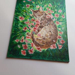 Puede incluir: Una pintura de un gato atigrado marr&oacute;n durmiendo en un campo de flores rosas. El gato est&aacute; enroscado en una bola con los ojos cerrados. Las flores est&aacute;n en flor y el fondo es un verde exuberante.