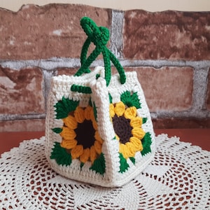 May include: A small, square-shaped crochet bag featuring a sunflower motif on each side. The bag is constructed with off-white yarn, showcasing yellow petals, dark brown centers, and green leaves. It includes two green crocheted handles at the top.