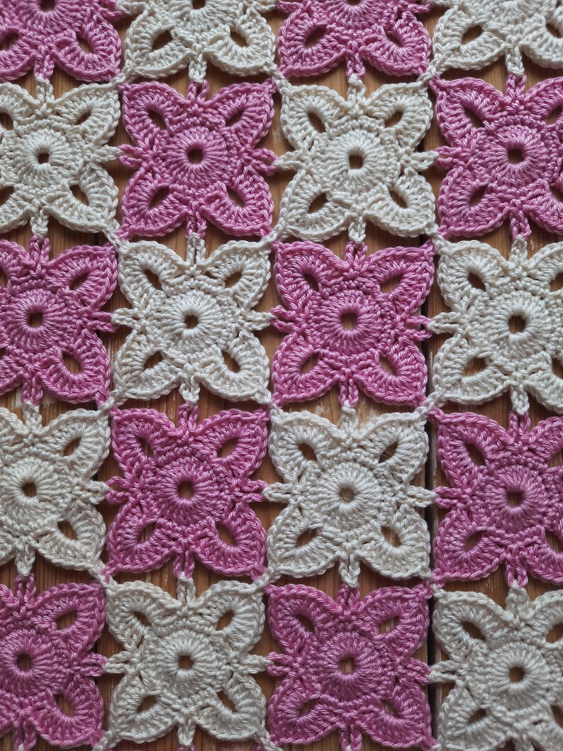 May include: A crocheted square pattern with alternating pink and cream coloured squares. The squares are made with a simple flower design.
