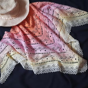 May include: A handmade, crochet shawl in a gradient of colors, from pale yellow to peach and pink, with a delicate, lacy border. A straw hat with a ribbon sits beside the shawl. The shawl is a beautiful accessory.